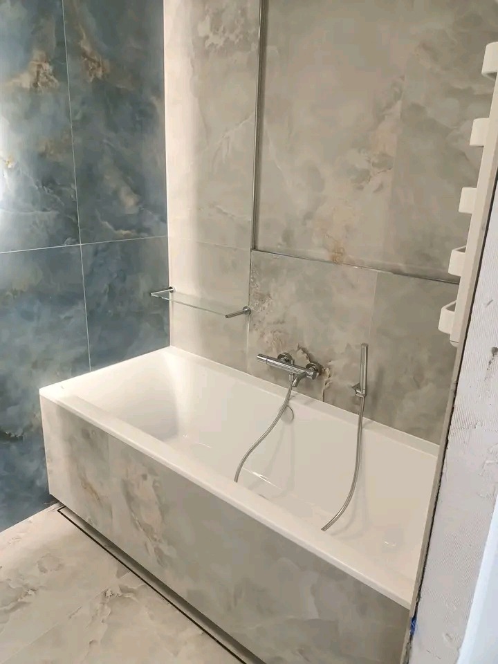 Wykończona łazienka z wanną obudowaną płytkami imitującymi marmur, armaturą chromowaną i grzejnikiem drabinkowym. Płytki ścienne w odcieniach szarości i błękitu.