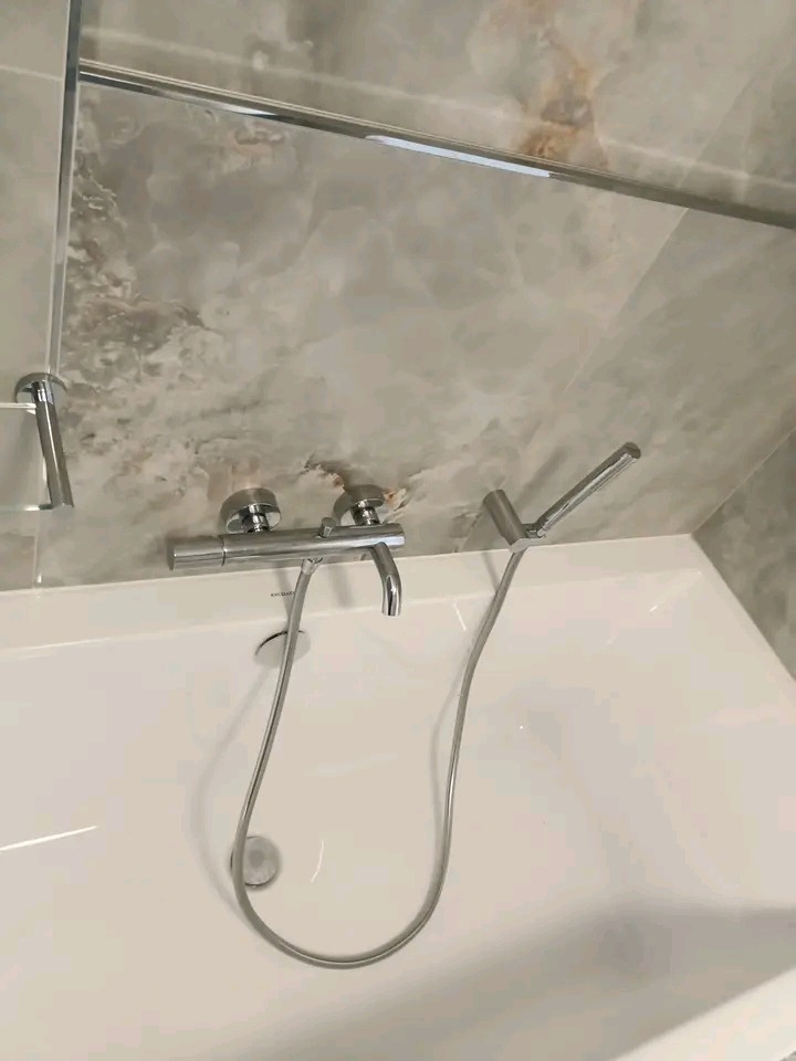 Biała wanna z chromowaną armaturą i deszczownicą na tle ściany wyłożonej płytkami imitującymi marmur w odcieniach szarości i beżu. Minimalistyczny design.