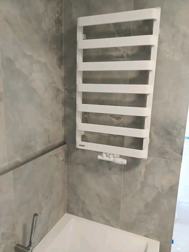 Biały grzejnik drabinkowy na szaro-beżowej ścianie z płytek imitujących marmur w nowoczesnej łazience. Widoczny fragment wanny i bateria.