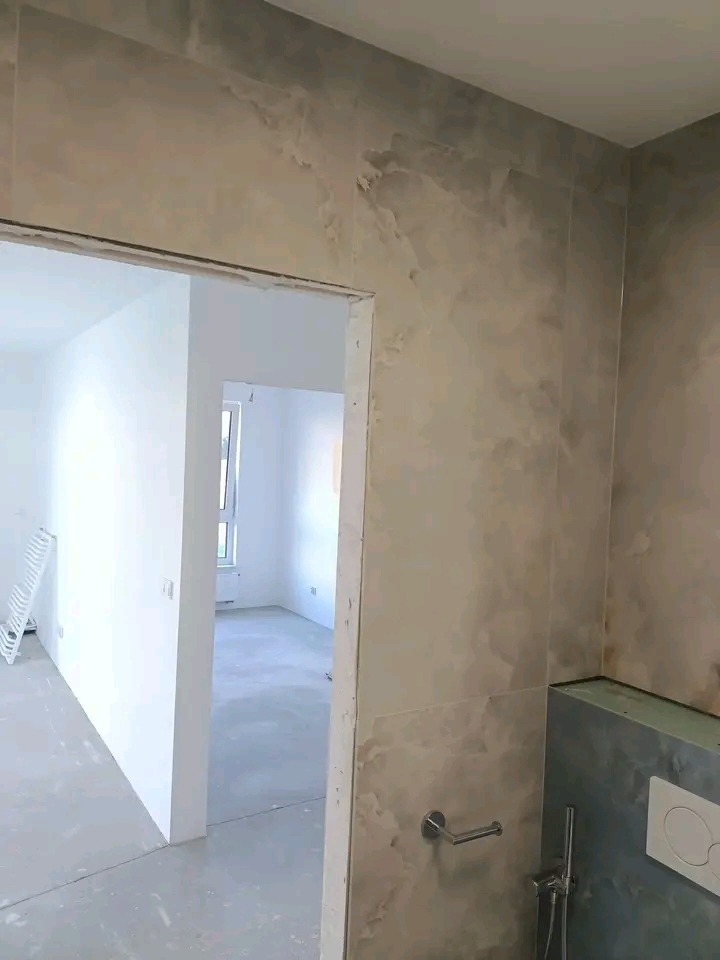 Wykończona łazienka z marmurowymi płytkami na ścianach i widokiem na sąsiednie, jeszcze nie wykończone pomieszczenie. Widoczny uchwyt na papier toaletowy.