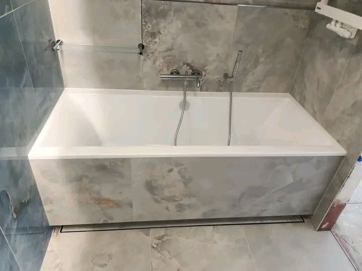 Nowoczesna, wykończona łazienka: wanna obudowana płytkami imitującymi marmur, szklana półka i armatura w kolorze srebrnym. Minimalistyczny design z dbałością o detale.