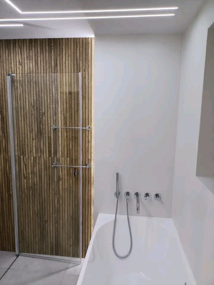 Nowoczesna łazienka z drewnianą ścianą, kabiną prysznicową ze srebrnymi okuciami i białą wanną z chromowaną armaturą. Minimalistyczne oświetlenie sufitowe.