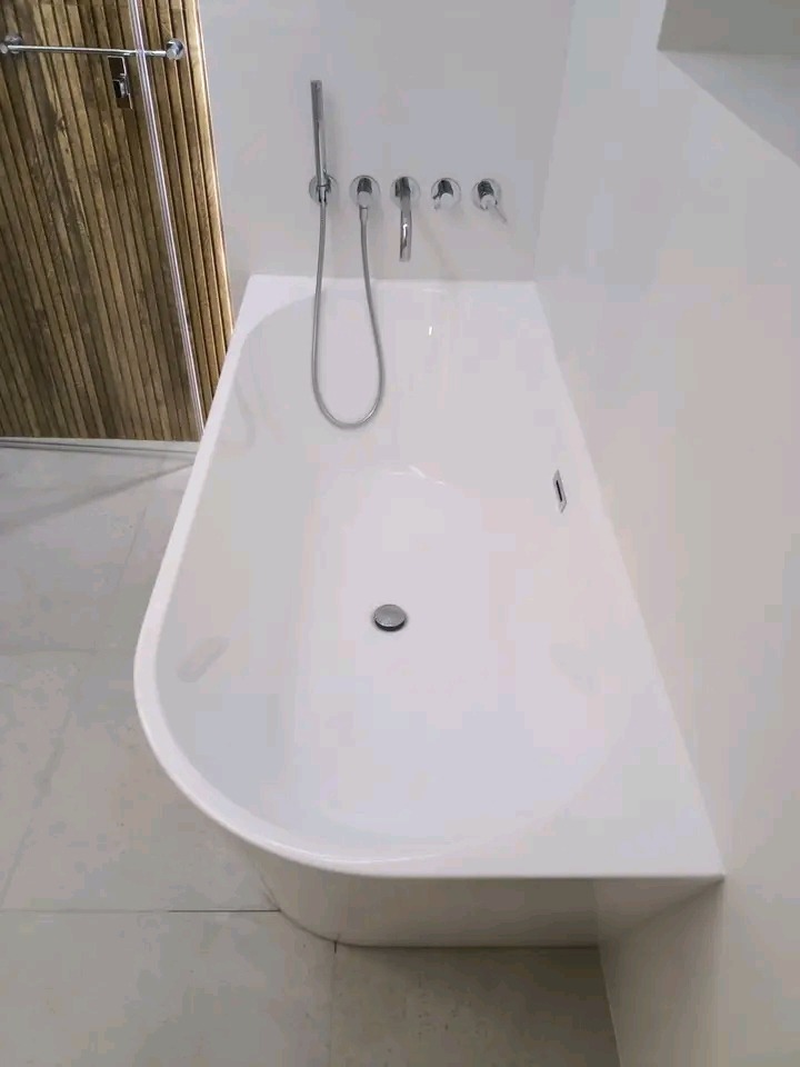 Biała, nowoczesna wanna wolnostojąca z chromowaną armaturą i prysznicem na tle ściany wykończonej panelami imitującymi drewno. Minimalistyczny design łazienki.