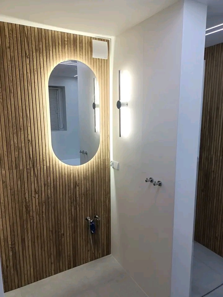 Nowoczesna łazienka z drewnianą okładziną ścienną, podświetlanym lustrem i minimalistycznym oświetleniem. Widoczne chromowane baterie i akcesoria.