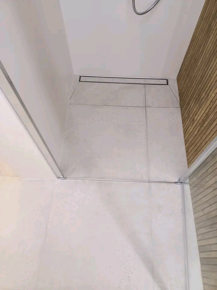 Nowoczesny odpływ liniowy w kabinie prysznicowej z jasnymi, kwadratowymi płytkami i drewnianą ścianą w tle. Minimalistyczny design z dbałością o detale.