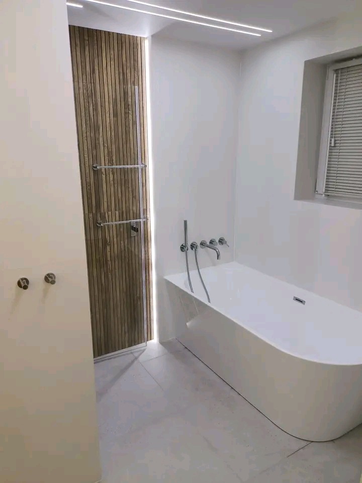 Nowoczesna łazienka z drewnianą ścianką działową i podświetleniem LED, białą wanną wolnostojącą i szarą marmurową podłogą, minimalistyczny design.