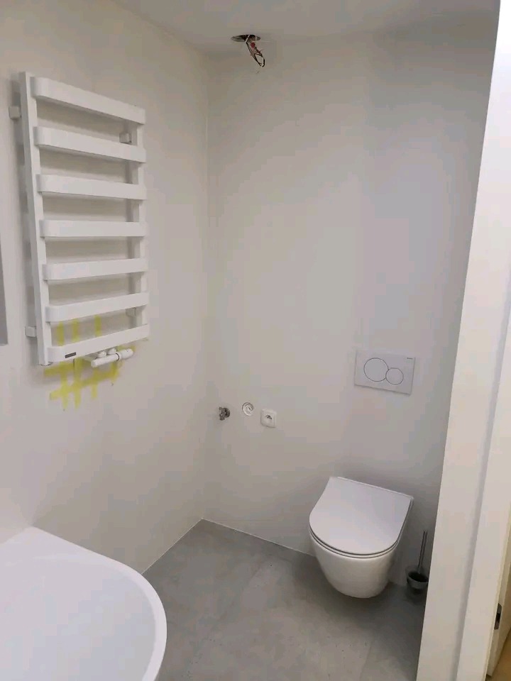 Nowoczesna, jasna łazienka z grzejnikiem drabinkowym, wiszącą toaletą i częściowo widoczną wanną. Widoczne instalacje elektryczne.