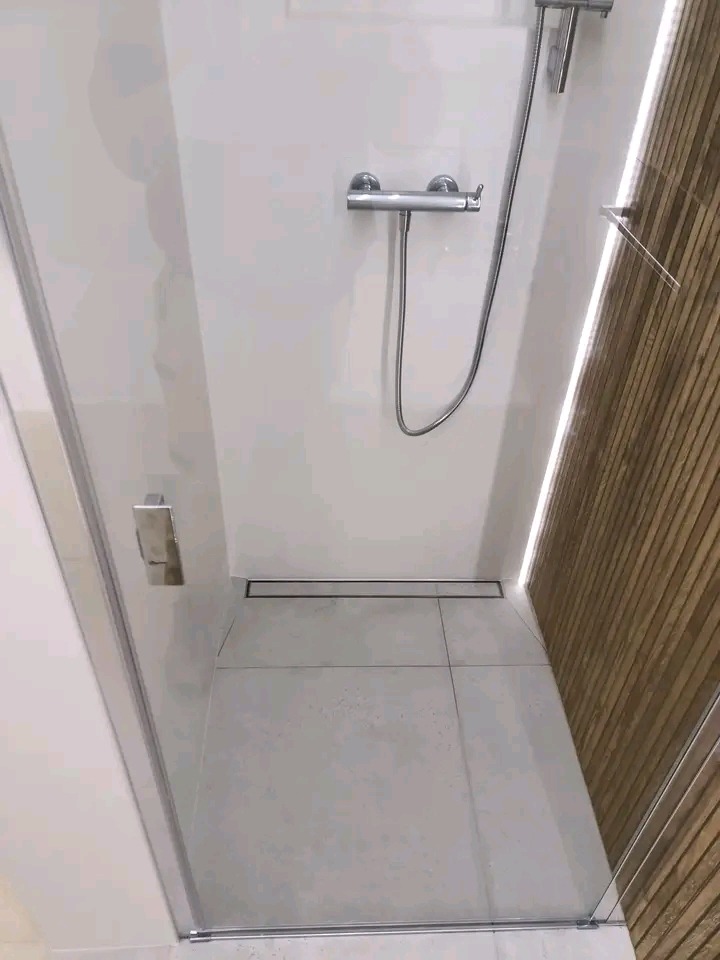 Nowoczesna kabina prysznicowa z odpływem liniowym, wykończona jasnymi płytkami i drewnianą ścianą. Widoczna armatura i szklane drzwi.