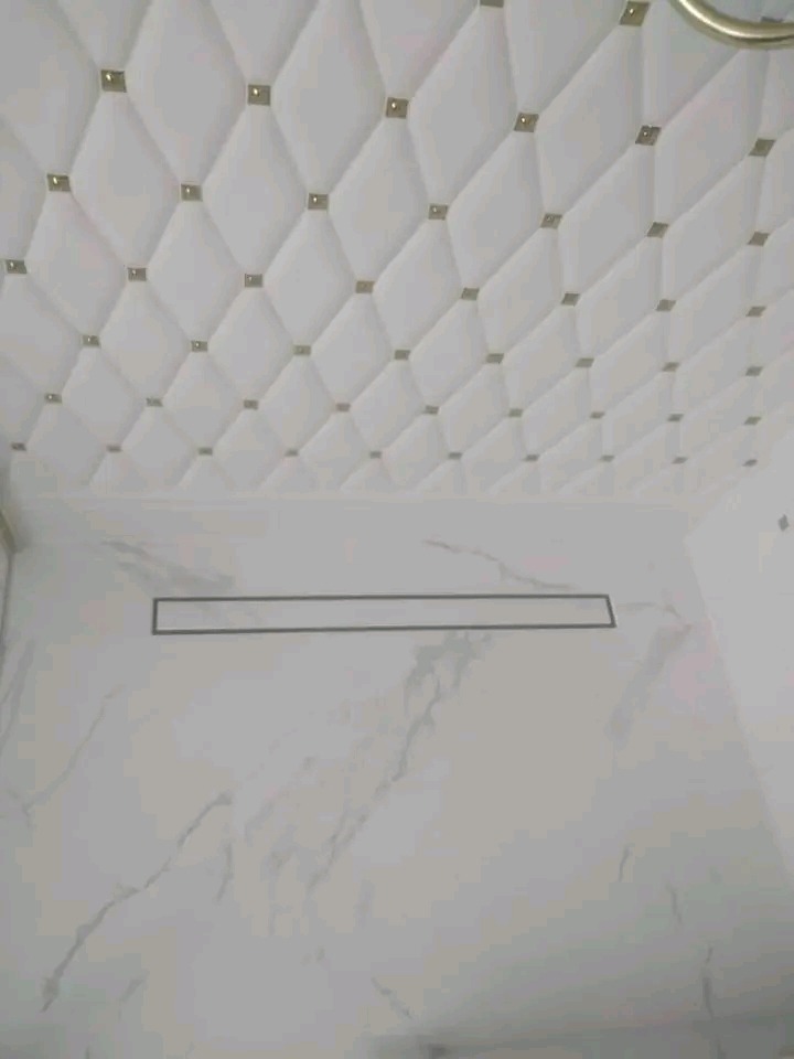 Wykończona łazienka z białą marmurową ścianą i sufitem z pikowanej skóry ekologicznej z ozdobnymi kwadratami. Widoczny odpływ liniowy.