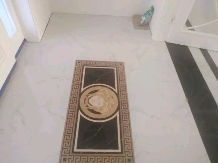 Biała marmurowa podłoga z czarnym, prostokątnym wzorem w stylu Versace, umieszczonym centralnie, oraz czarne pasy przy drzwiach po prawej stronie.