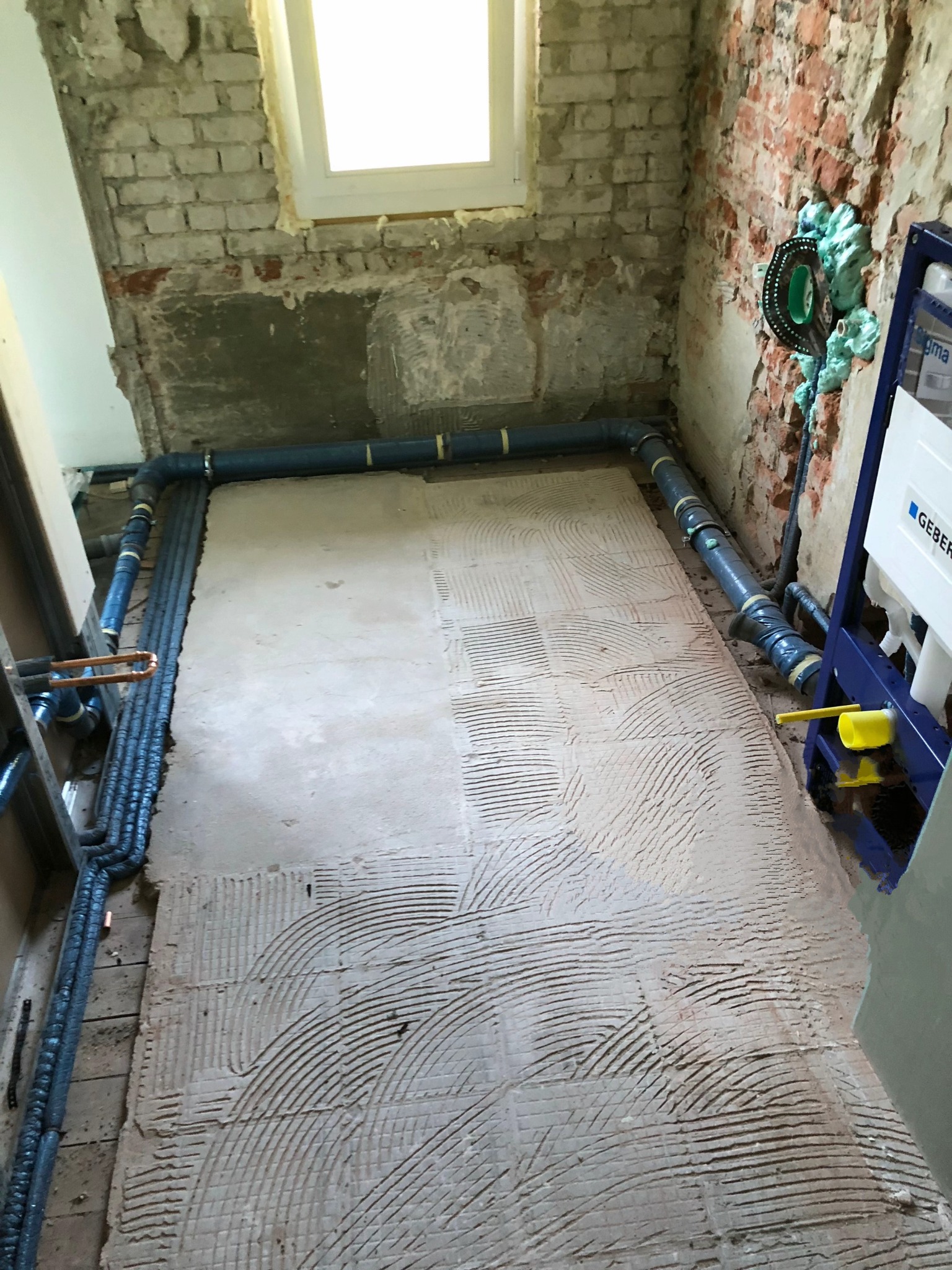 Przygotowanie podłoża pod płytki w łazience: na podłodze widoczna warstwa kleju z charakterystycznymi rowkami, rury instalacyjne i stelaż podtynkowy Geberit.