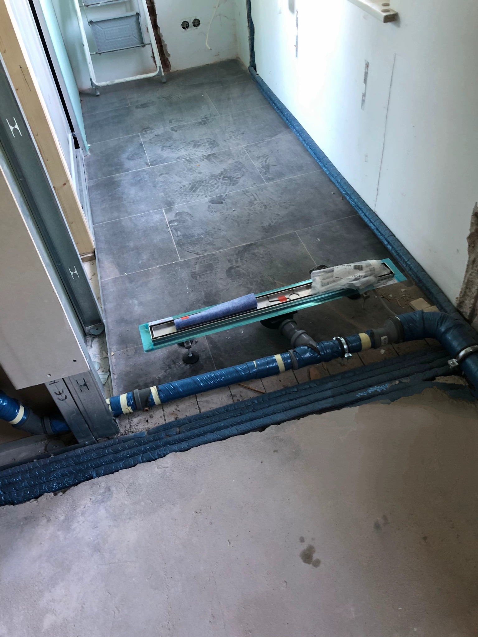 Ułożona szara terakota w łazience z zamontowanym odpływem liniowym i rurami kanalizacyjnymi. Widoczne ślady po butach na płytkach.