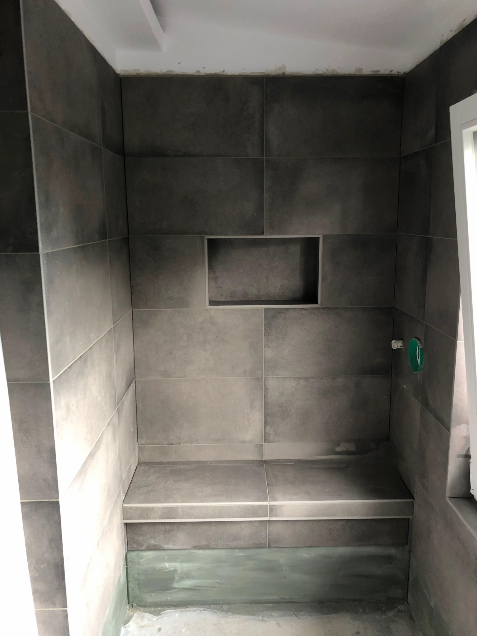 Wykończona wnęka prysznicowa z siedziskiem, wyłożona szarymi płytkami w dużym formacie. Widoczna wnęka w ścianie i fragment okna po prawej stronie.