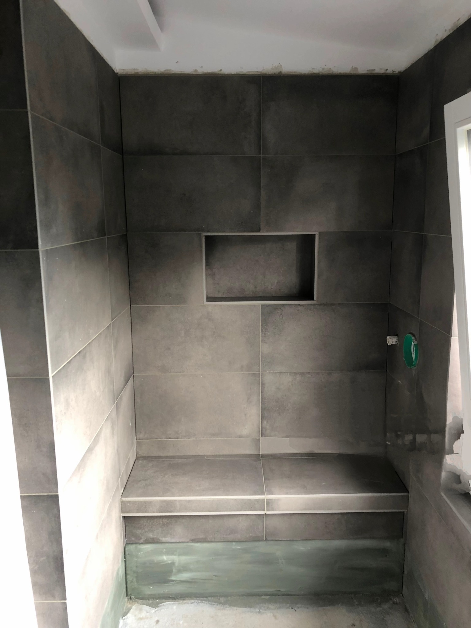 Wykończona wnęka prysznicowa z siedziskiem, wyłożona szarymi płytkami. Widoczna wnęka w ścianie i fragment instalacji. Minimalistyczny design.