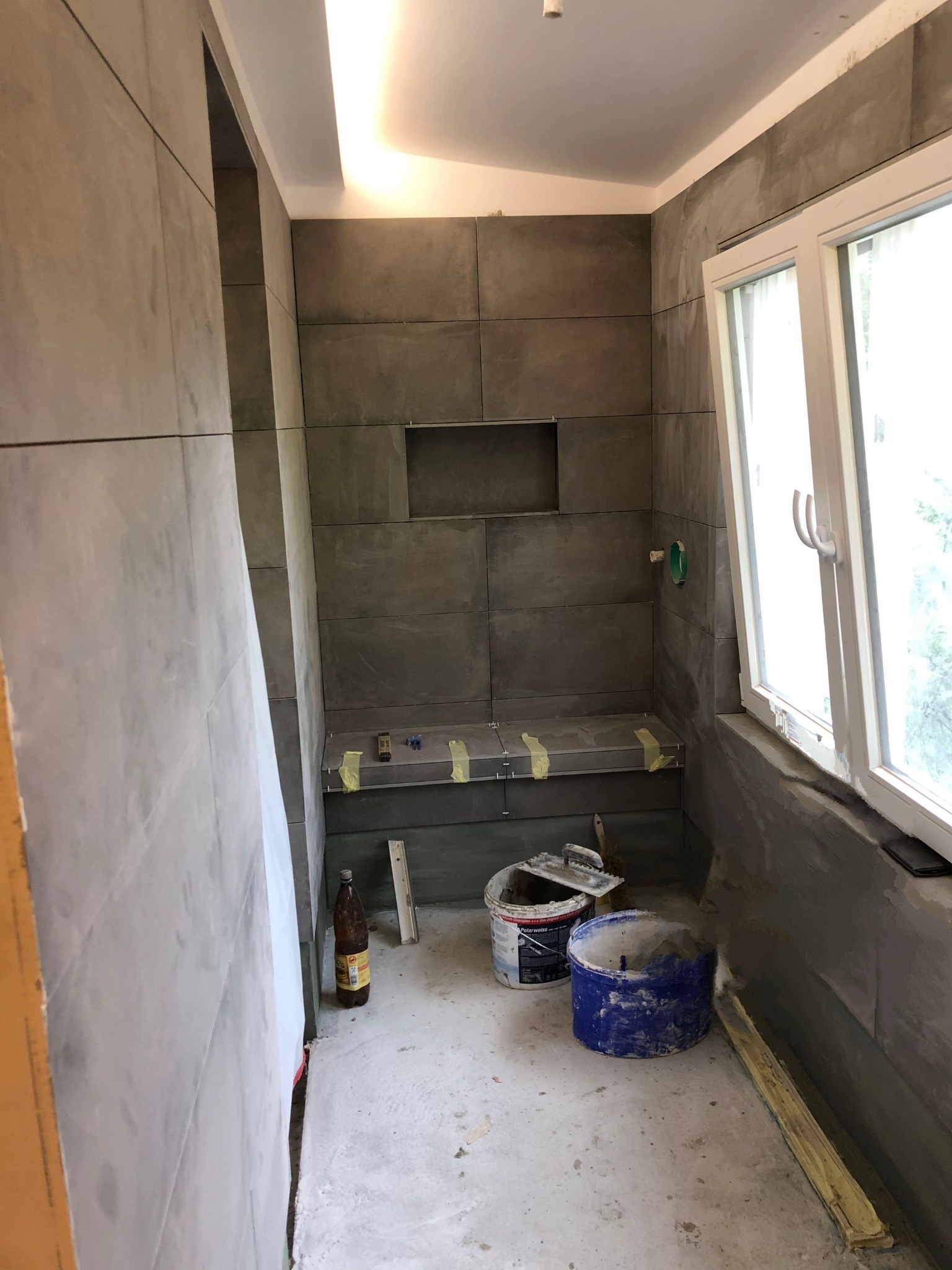 Łazienka w trakcie wykańczania: szare płytki na ścianach, widoczne narzędzia glazurnicze, okno z białymi ramami. Etap prac wykończeniowych, surowy stan podłogi.