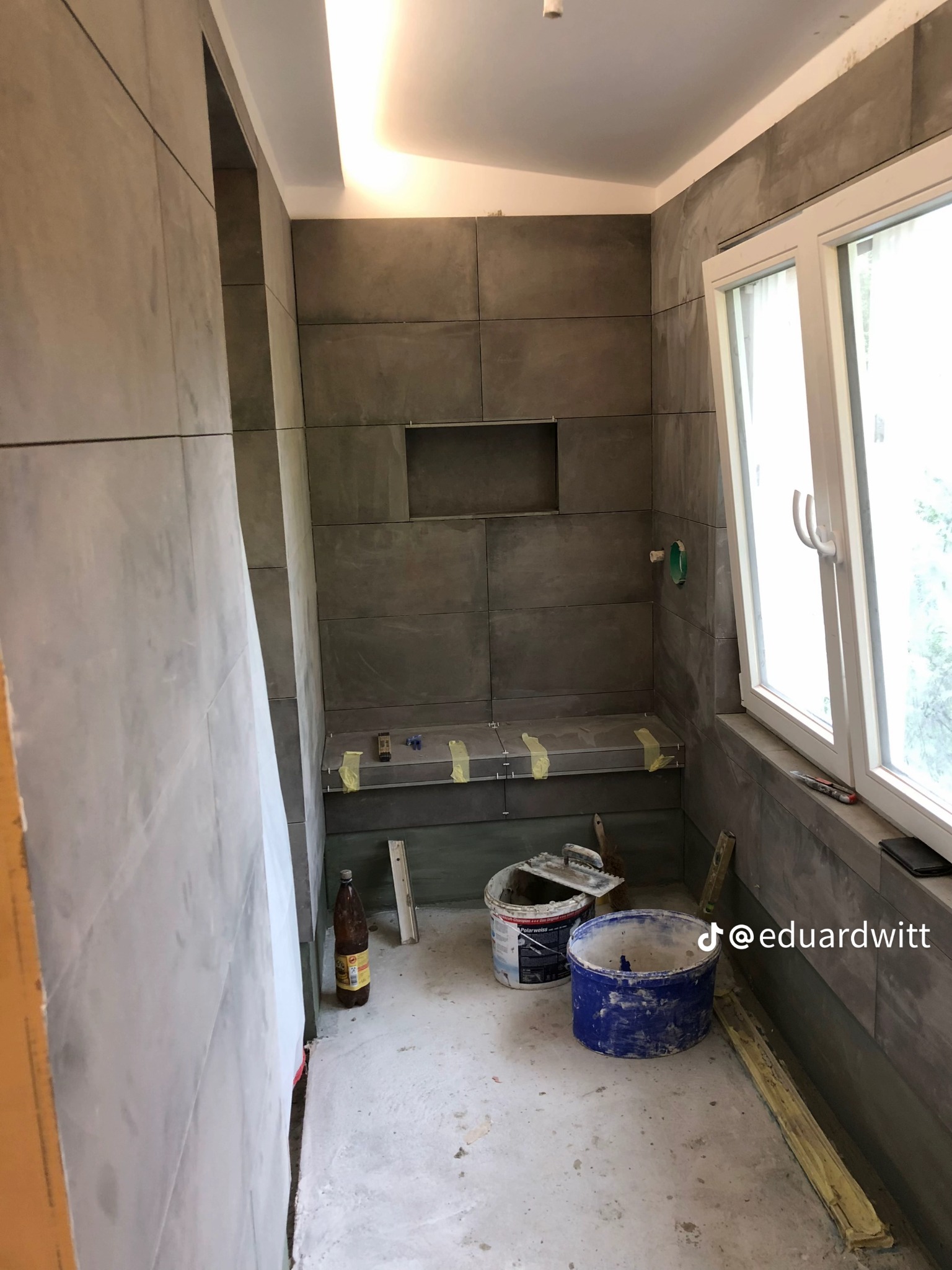 Łazienka w trakcie wykańczania: szare płytki wielkoformatowe na ścianach, widoczne narzędzia i wiaderka, okno z widokiem na zieleń. Minimalistyczny design w surowym wydaniu.