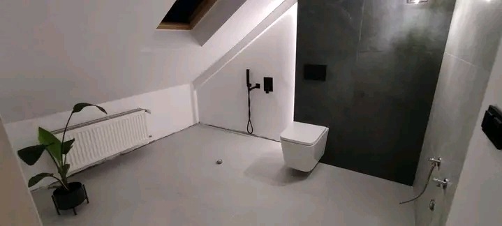 Nowoczesna łazienka na poddaszu z czarną armaturą i szarą podłogą żywiczną. Uwagę przykuwa minimalistyczna toaleta podwieszana i roślina doniczkowa.