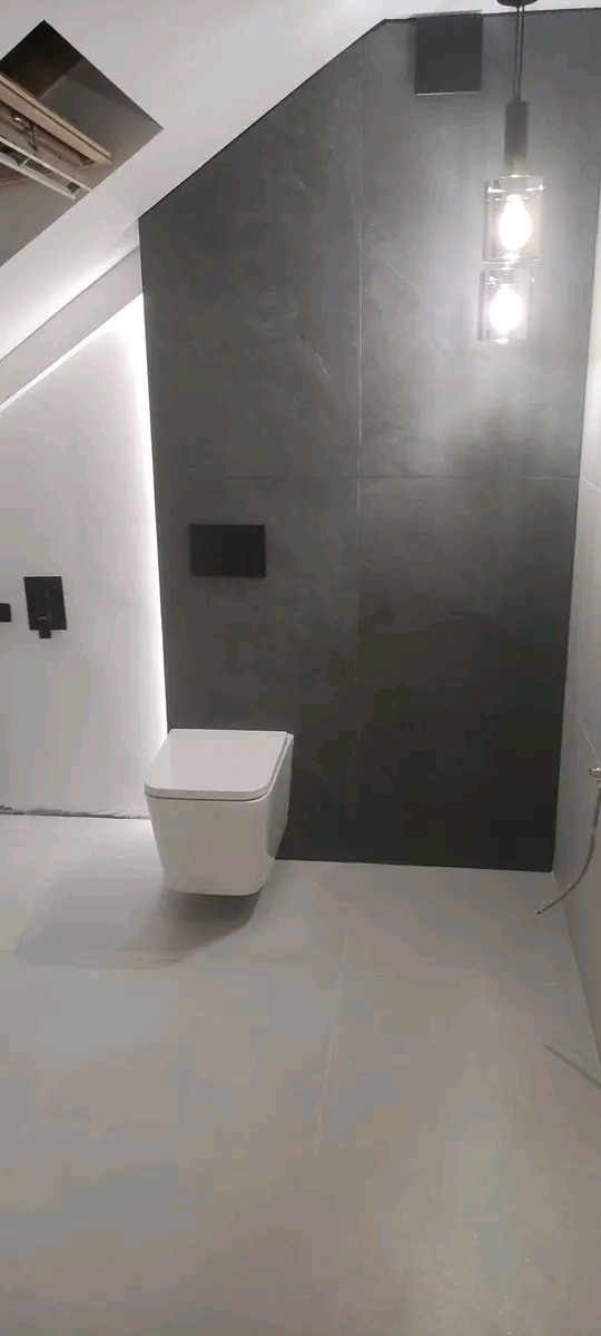 Nowoczesna łazienka z wiszącą toaletą, szarą ścianą z płytek i minimalistycznym oświetleniem. Uwagę zwraca nietypowe ułożenie płytek na ścianie i sufit ze skosem.