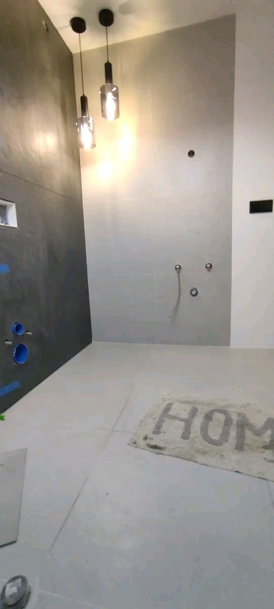 Nowoczesna łazienka w trakcie wykańczania: szare płytki, minimalistyczne lampy wiszące, przygotowane instalacje wodne i dywanik z napisem 'HOME'.