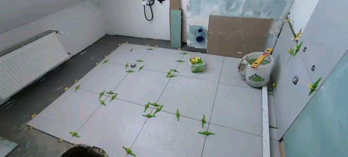 Jasne płytki podłogowe w trakcie układania w łazience na poddaszu, z użyciem zielonych krzyżyków dystansowych. Widoczny kubeł z zaprawą i poziomica.