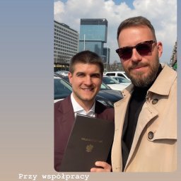 PROSPECT R.E.INVEST SPÓŁKA Z OGRANICZONĄ ODPOWIEDZIALNOŚCIĄ - Uśmiechnięci mężczyźni po podpisaniu umowy sprzedaży nieruchomości. Jeden trzyma Akt Notarialny. W tle budynki miejskie i samochody.