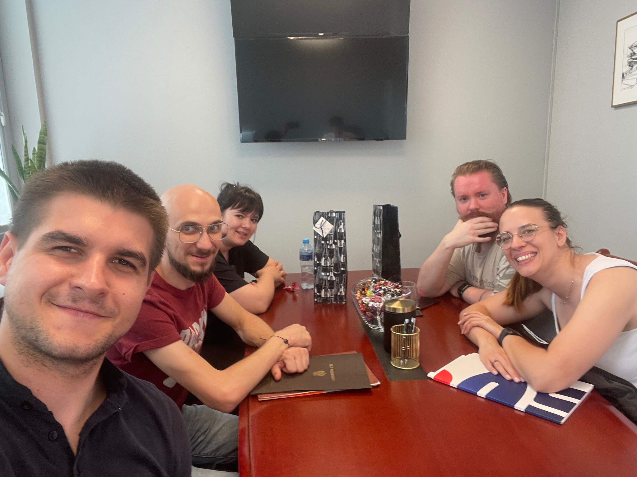 Zespół w biurze siedzi przy czerwonym stole. Na stole dokumenty, słodycze, w tle telewizor i roślina doniczkowa. Uśmiechnięte twarze, pozytywna atmosfera.