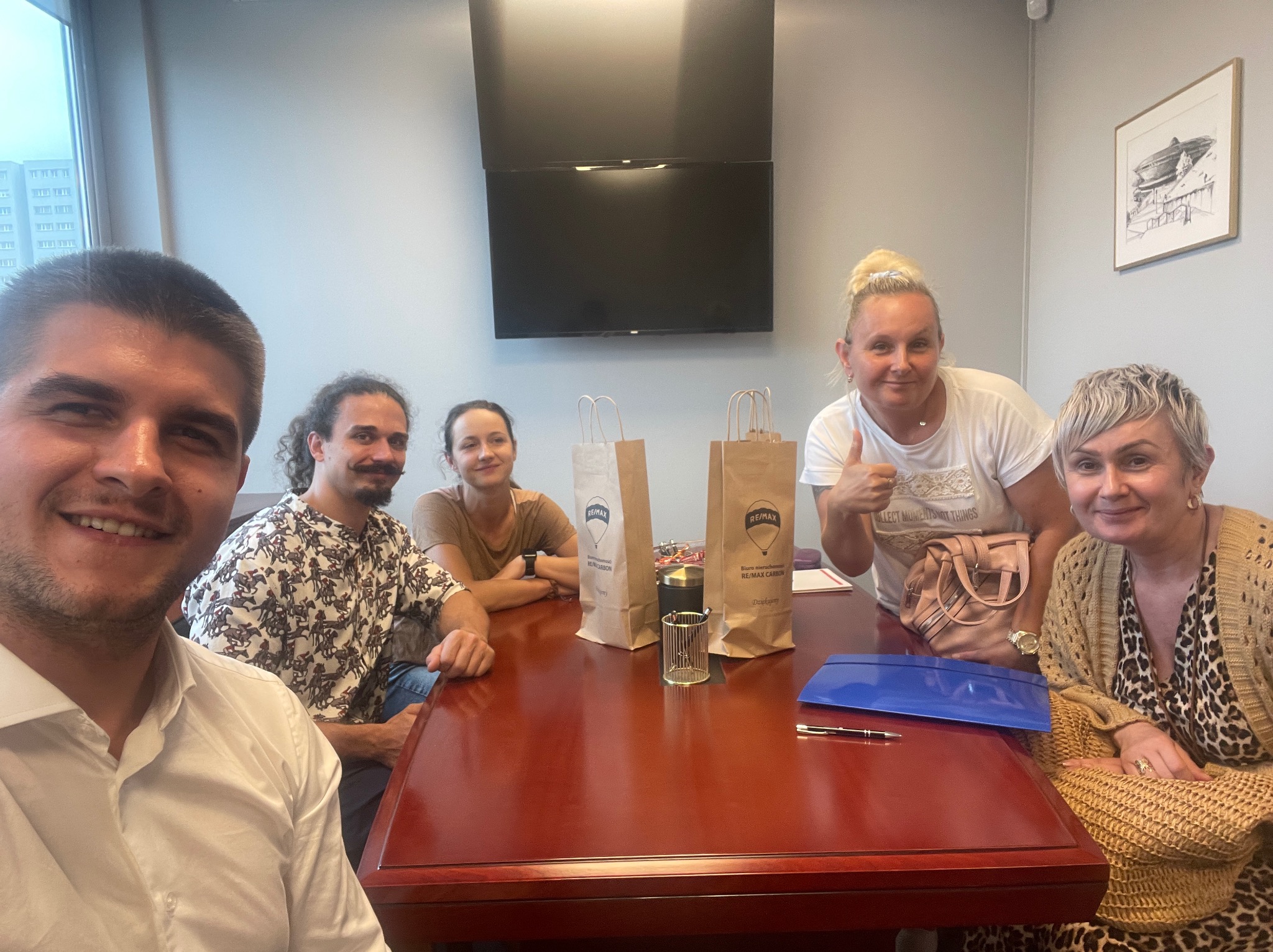 Grupa osób przy stole konferencyjnym z torbami RE/MAX, uśmiechnięta blondynka pokazuje kciuk w górę, nowoczesne biuro z telewizorem i obrazem na ścianie.