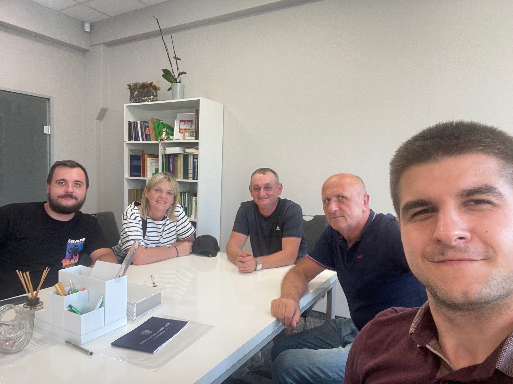 Kameralne spotkanie zespołu przy białym stole w biurze. Uśmiechnięte twarze, swobodna atmosfera, w tle regał z książkami. Selfie zrobione przez jednego z uczestników.