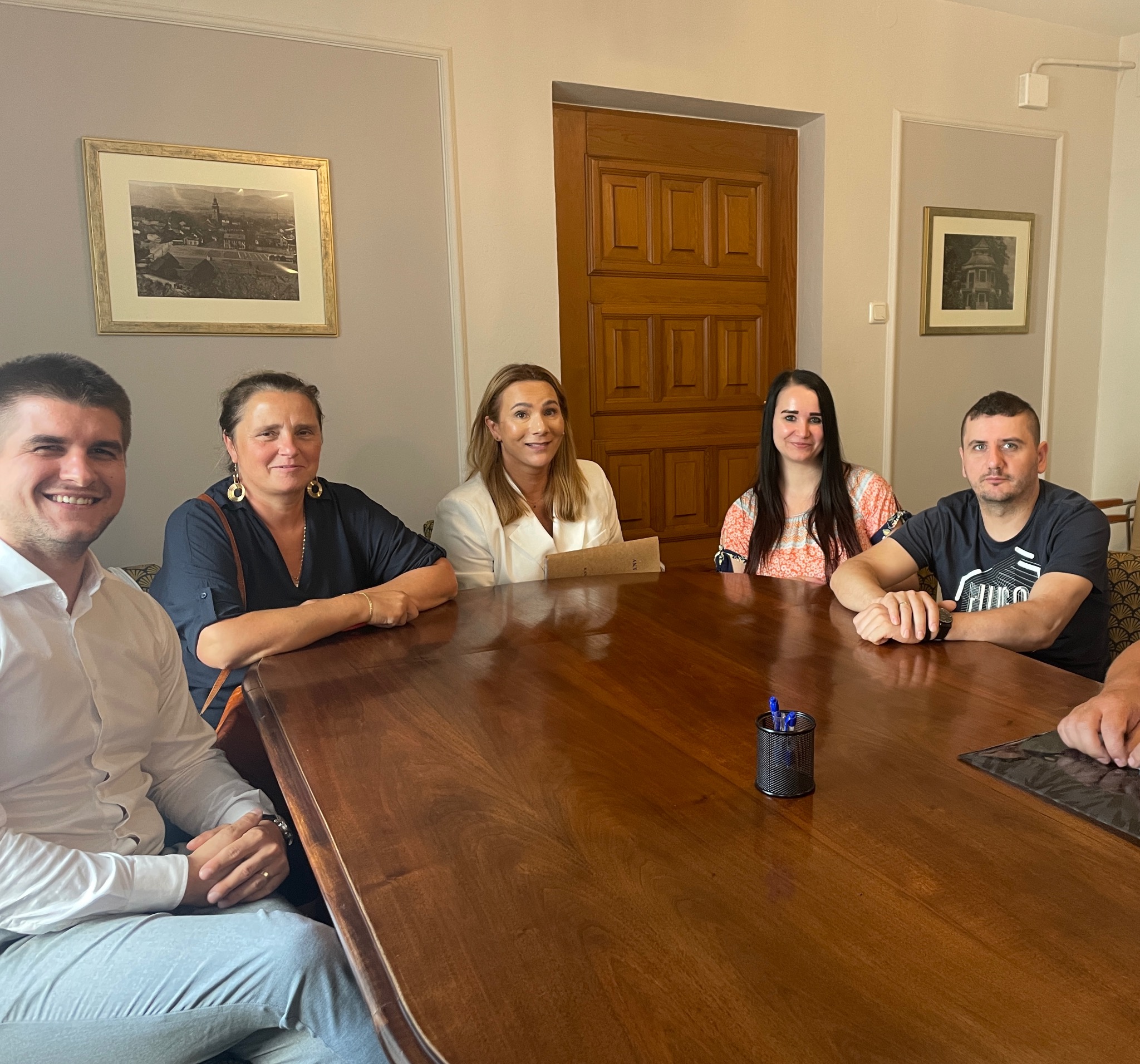 Grupa osób siedzi przy dużym drewnianym stole w jasnym pomieszczeniu z obrazami na ścianach i drewnianymi drzwiami w tle. Uśmiechnięte twarze uczestników spotkania.