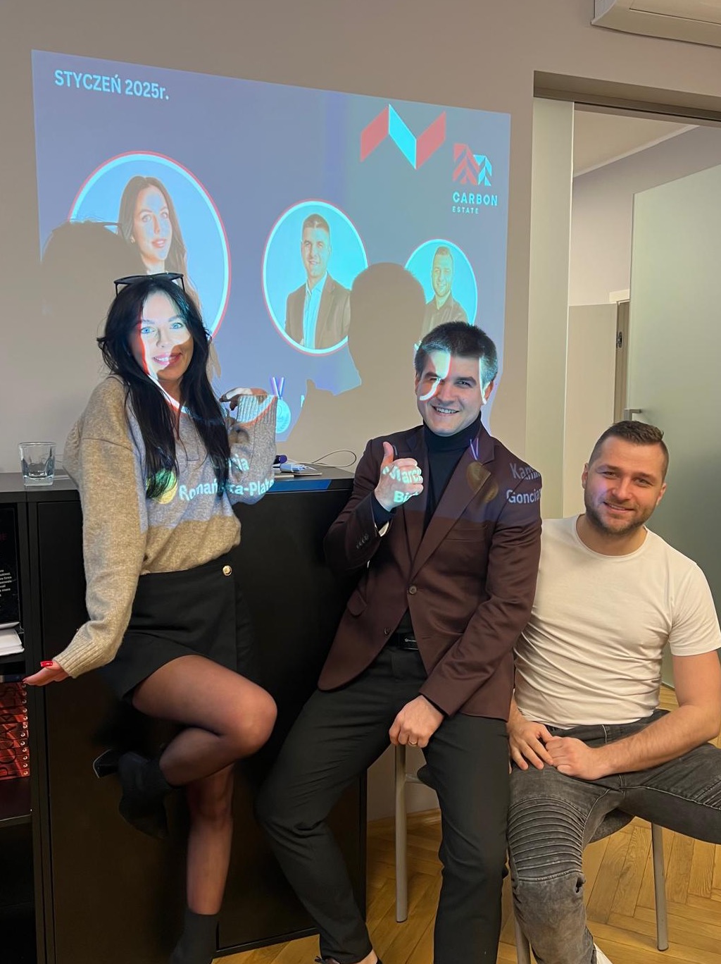 Trzy osoby pozują na tle prezentacji Carbon Estate ze zdjęciami pracowników i logotypem. Mężczyzna w bordowej marynarce pokazuje kciuk w górę.