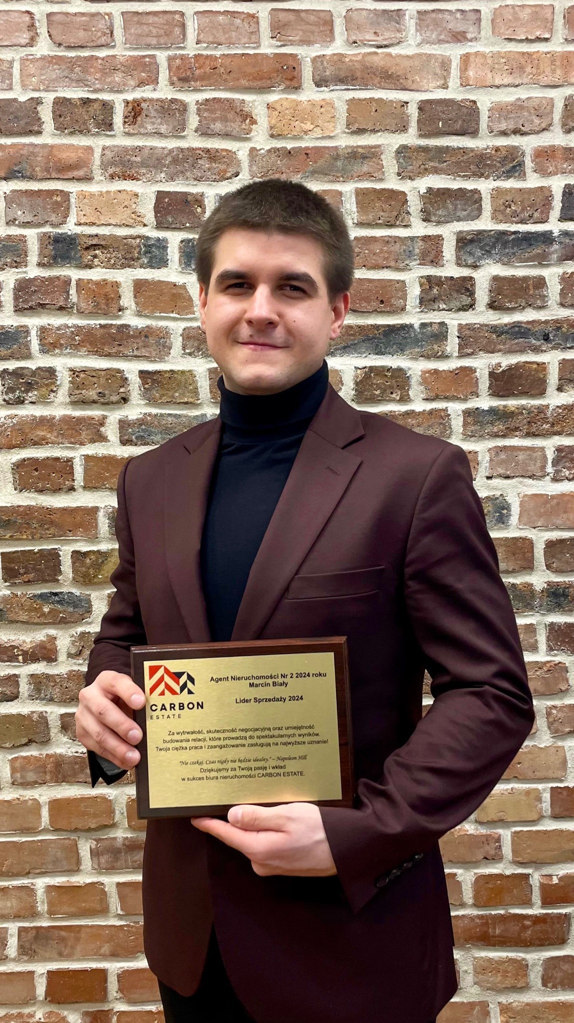 Agent nieruchomości Marcin Biały z nagrodą 'Lider Sprzedaży 2024' na tle ceglanej ściany. Profesjonalny wizerunek biura Carbon Estate.