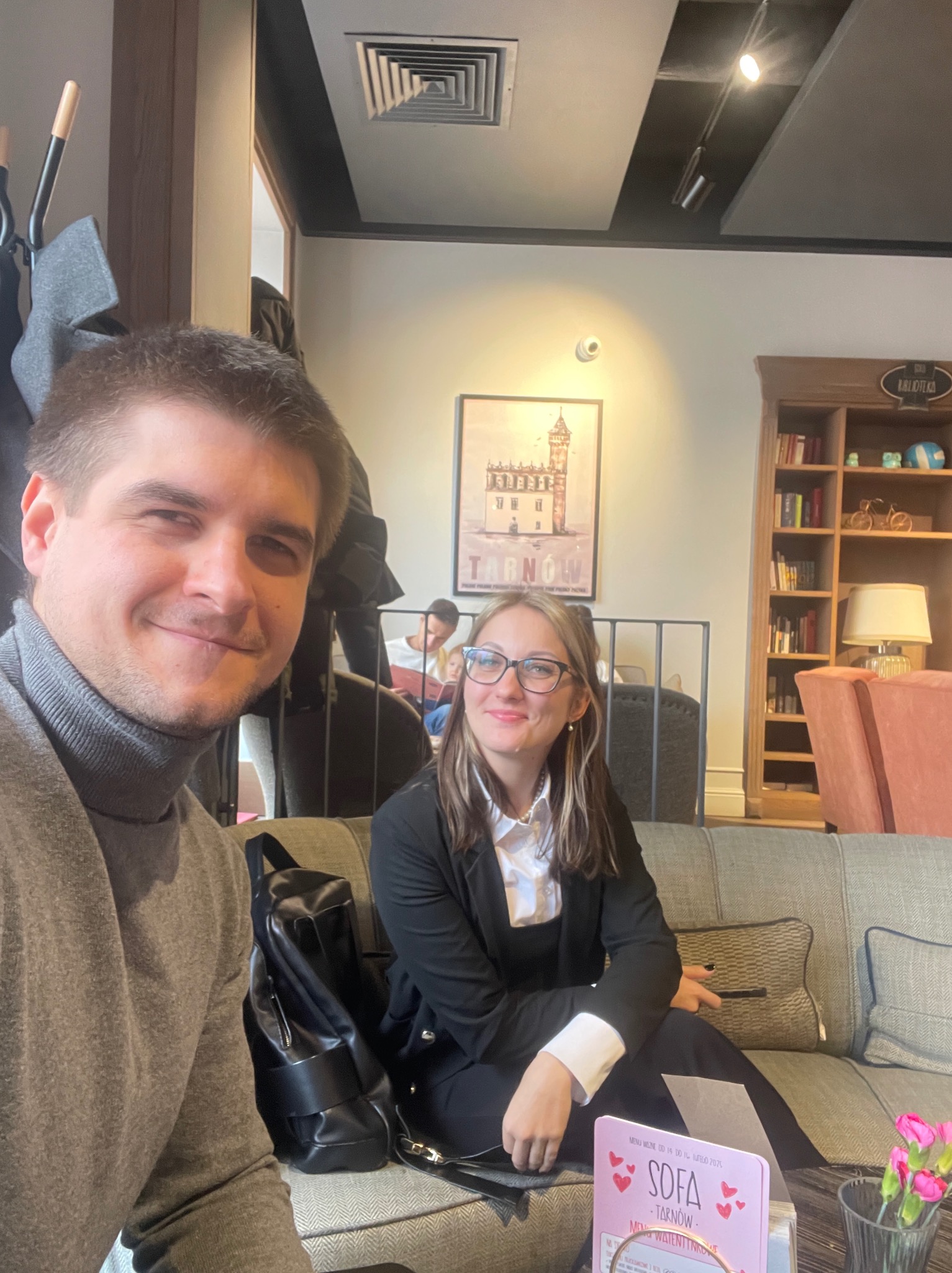 Dwoje uśmiechniętych ludzi siedzi na kanapie w kawiarni. W tle plakat z Tarnowem i regał z książkami. Mężczyzna w golfie, kobieta w marynarce.