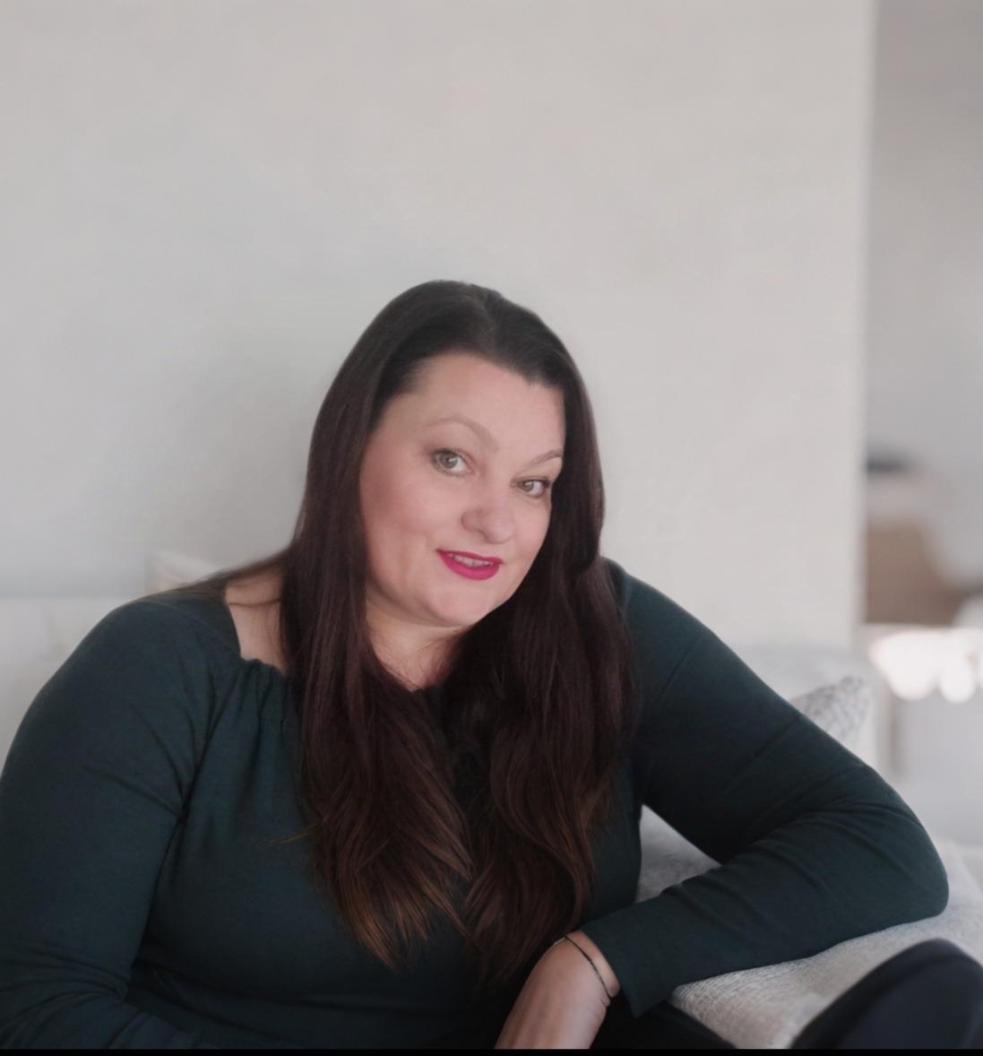 Portret uśmiechniętej kobiety z długimi, ciemnymi włosami siedzącej na jasnej kanapie w minimalistycznym wnętrzu.