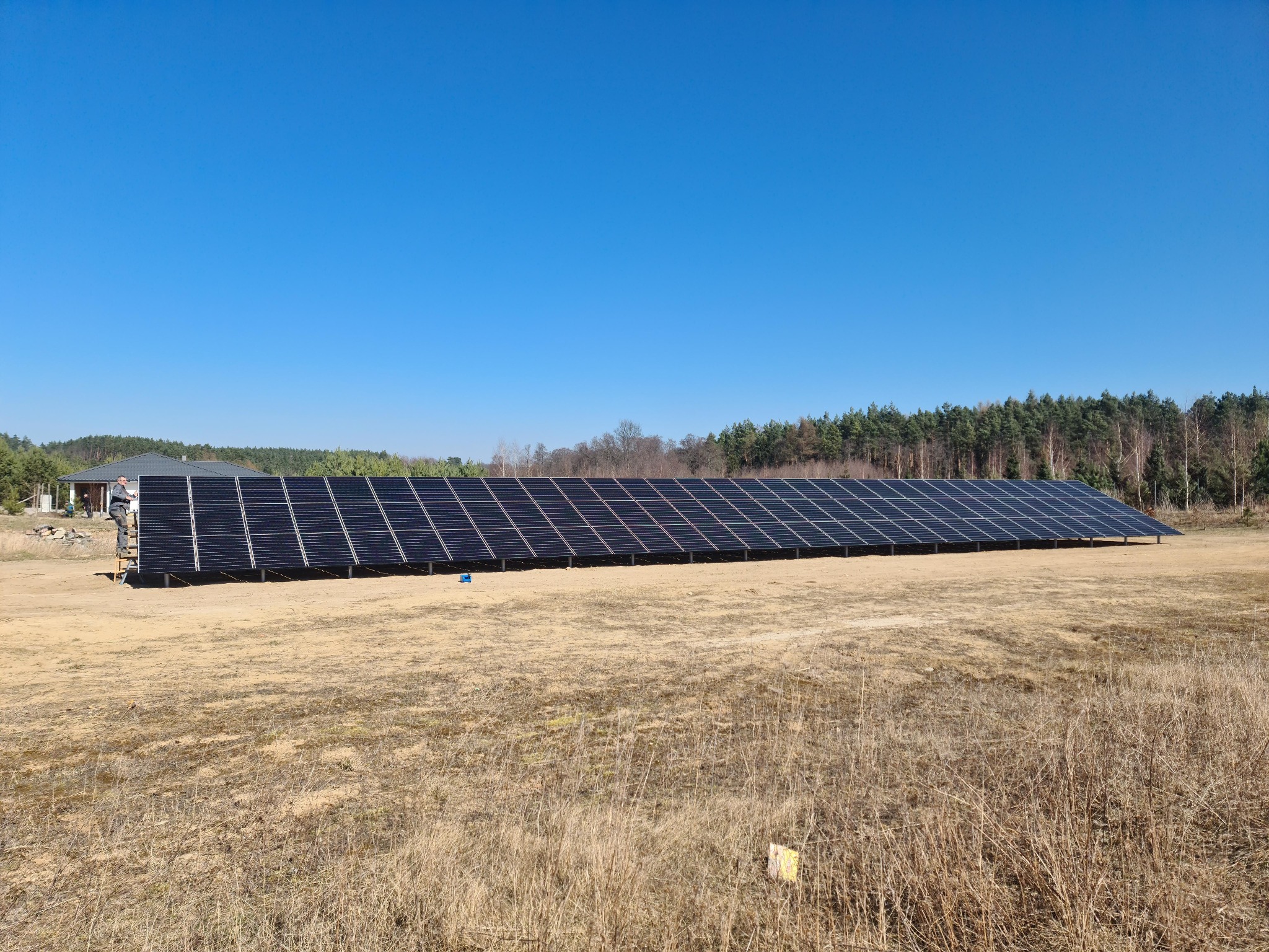 Rozległa instalacja paneli słonecznych na polu, z pracownikiem na drabinie przy jednym z końców. W tle las i dom, błękitne niebo.