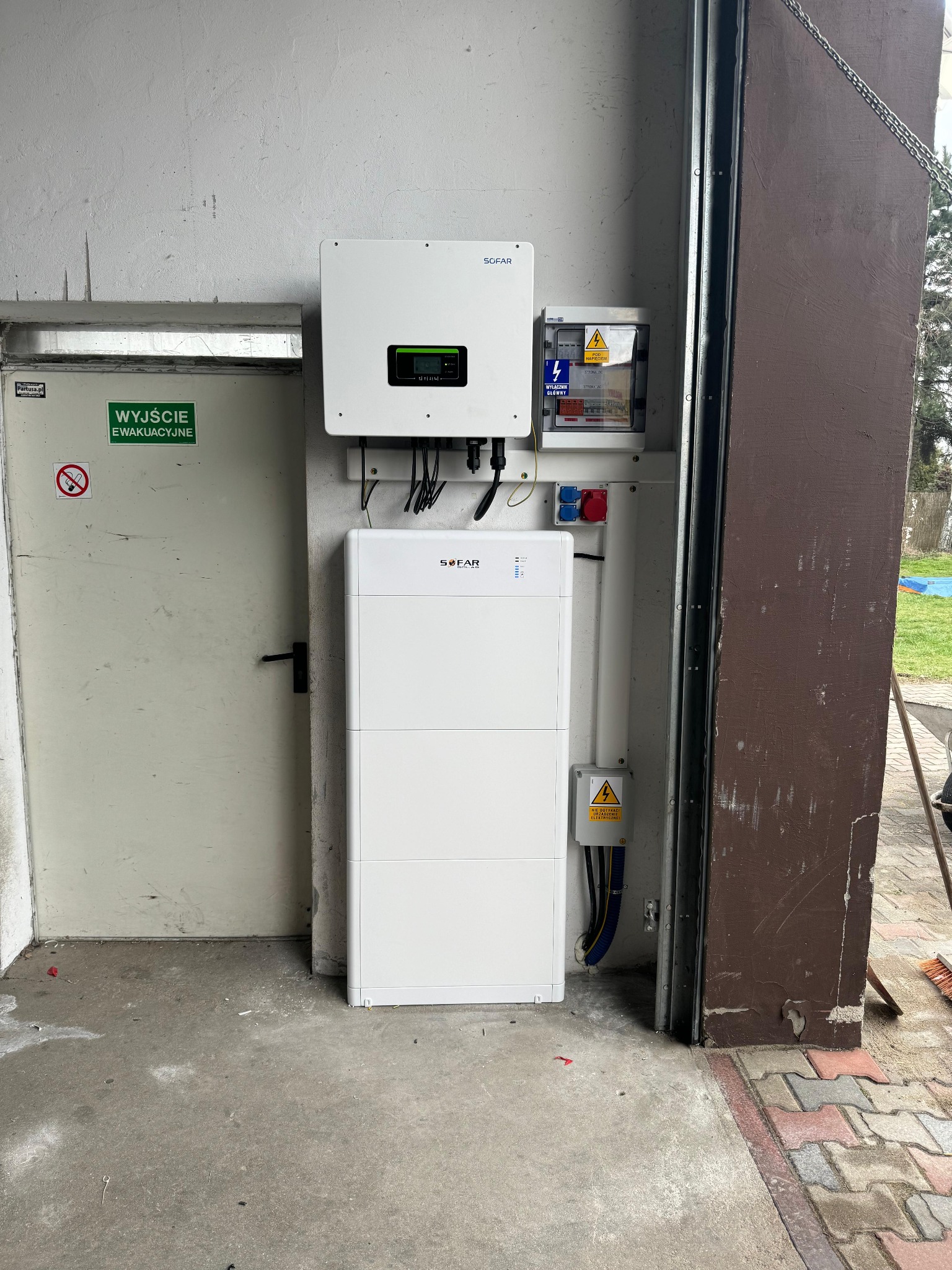 Biały inwerter SOFAR z akumulatorem energii, skrzynka elektryczna i gniazda siłowe na ścianie w garażu, obok drzwi ewakuacyjnych. Widoczne kable i przewody.
