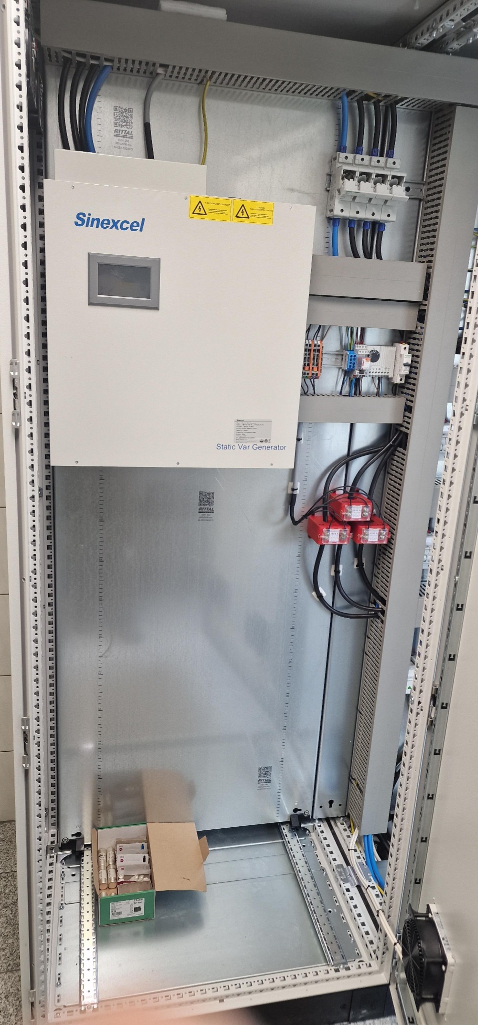 Wnętrze szafy elektrycznej z generatorem statycznym VAR Sinexcel, widoczne przewody, zabezpieczenia i wentylator. Elementy zamontowane na metalowej konstrukcji.
