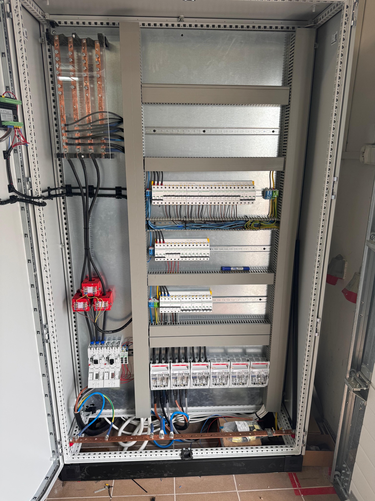 Otwarta szafa elektryczna z widocznymi elementami instalacji: kable, bezpieczniki, listwy zaciskowe. Widoczne przewody w kolorach: czarnym, niebieskim, żółto-zielonym.