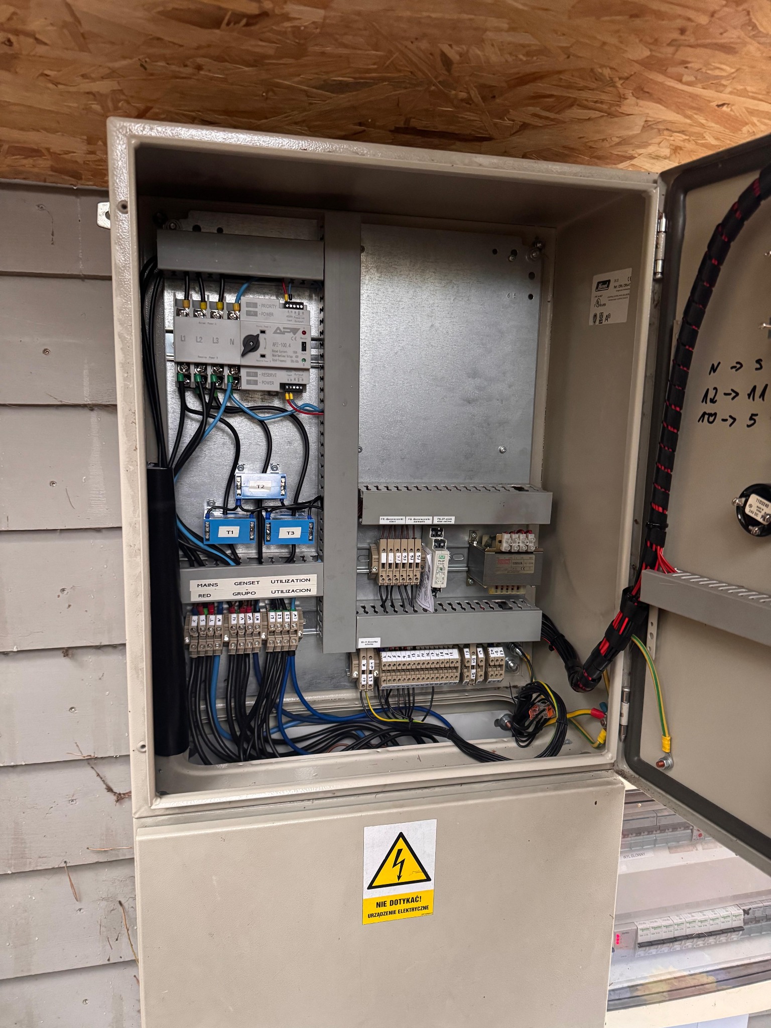 Otwarta skrzynka elektryczna z widocznymi przewodami, bezpiecznikami i elementami sterującymi. Żółty znak ostrzegawczy o napięciu. Widoczne oznaczenia i etykiety.