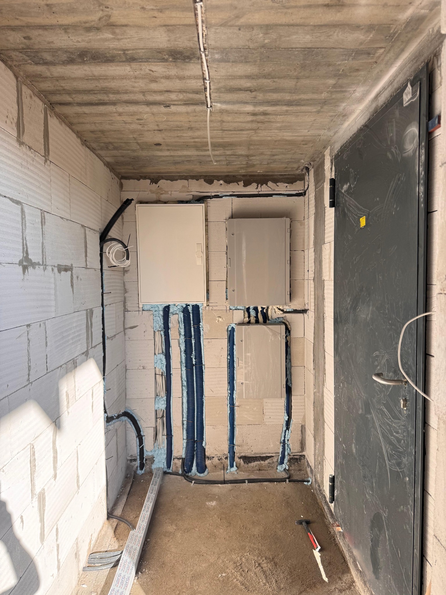 Wnętrze w trakcie prac elektrycznych: widoczne puszki elektryczne, kable w peszlach, ściany z betonu komórkowego i surowy betonowy strop. Częściowo widoczna metalowa ościeżnica.