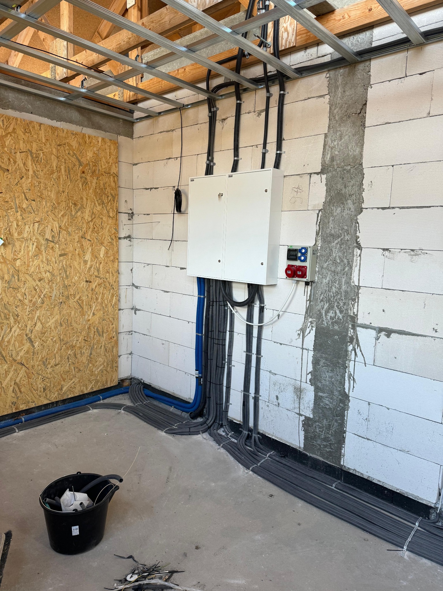 Rozdzielnia elektryczna z przewodami w peszlach, zamontowana na ścianie z betonu komórkowego w surowym wnętrzu. Widoczne kable i puszki instalacyjne.
