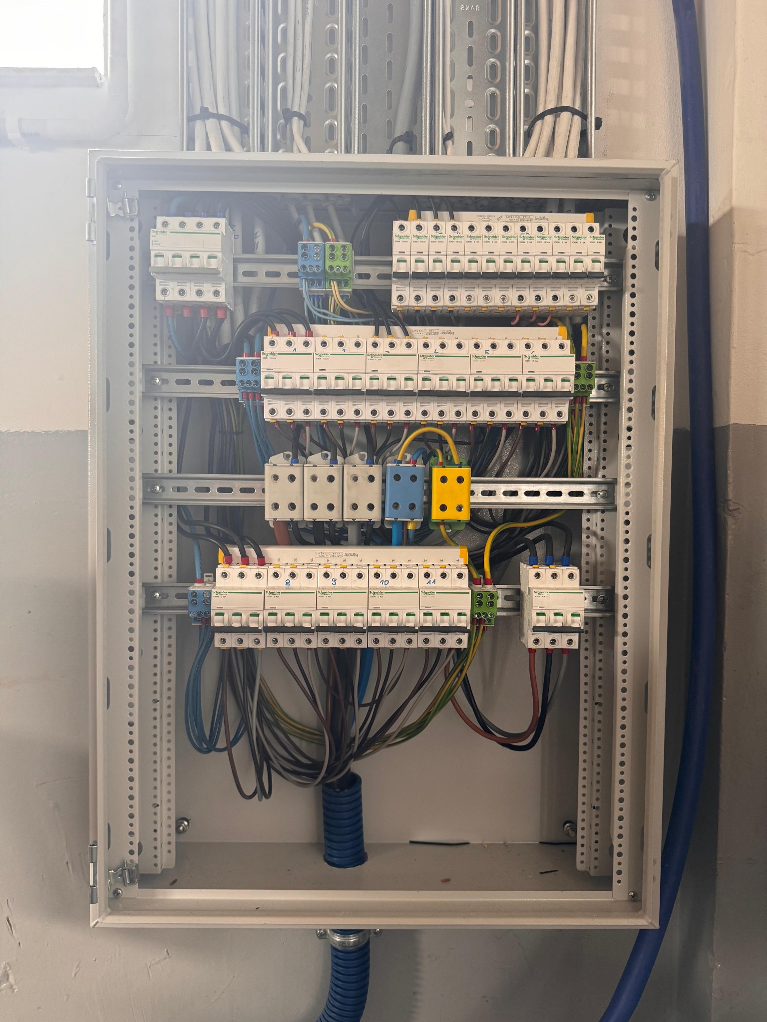 Rozdzielnica elektryczna Schneider z licznymi zabezpieczeniami, widoczne przewody w peszlu. Elementy zamontowane na szynach DIN wewnątrz metalowej obudowy.