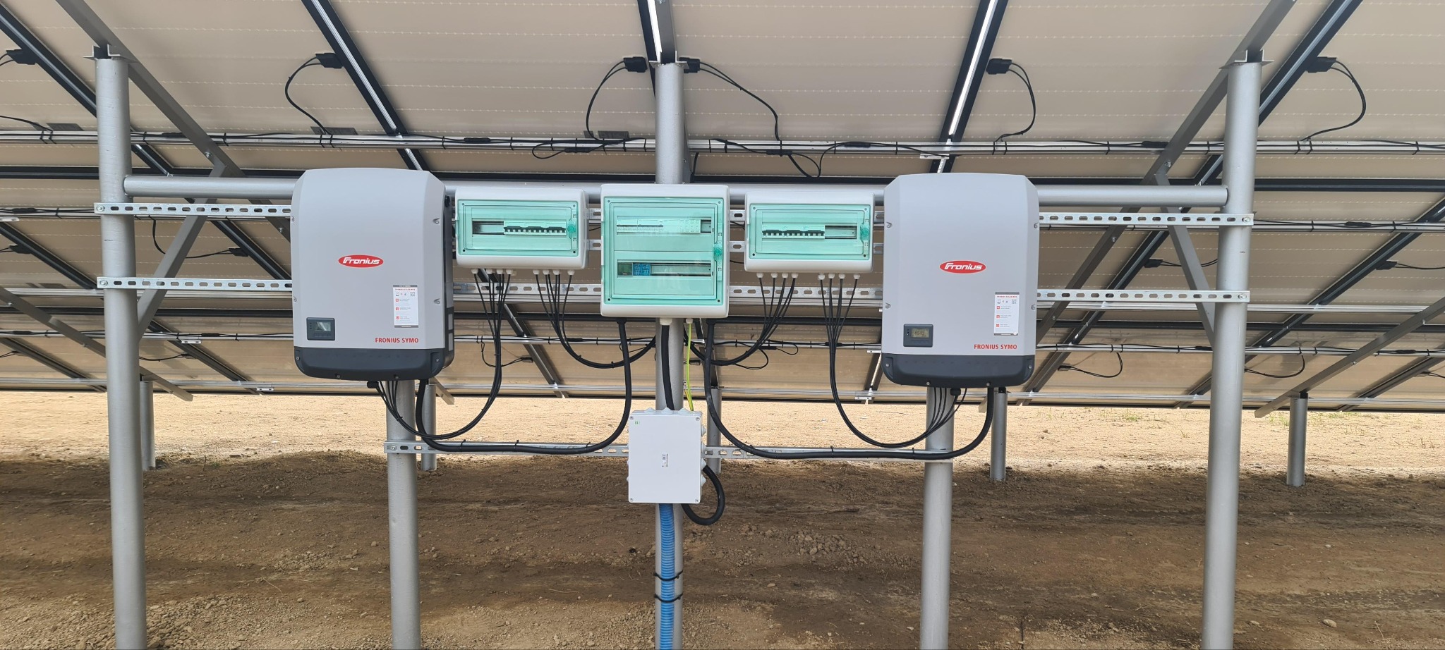 Instalacja paneli słonecznych z widocznymi inwerterami Fronius Symo i rozdzielnicami elektrycznymi, zamontowanymi na metalowej konstrukcji. Widok od spodu paneli.