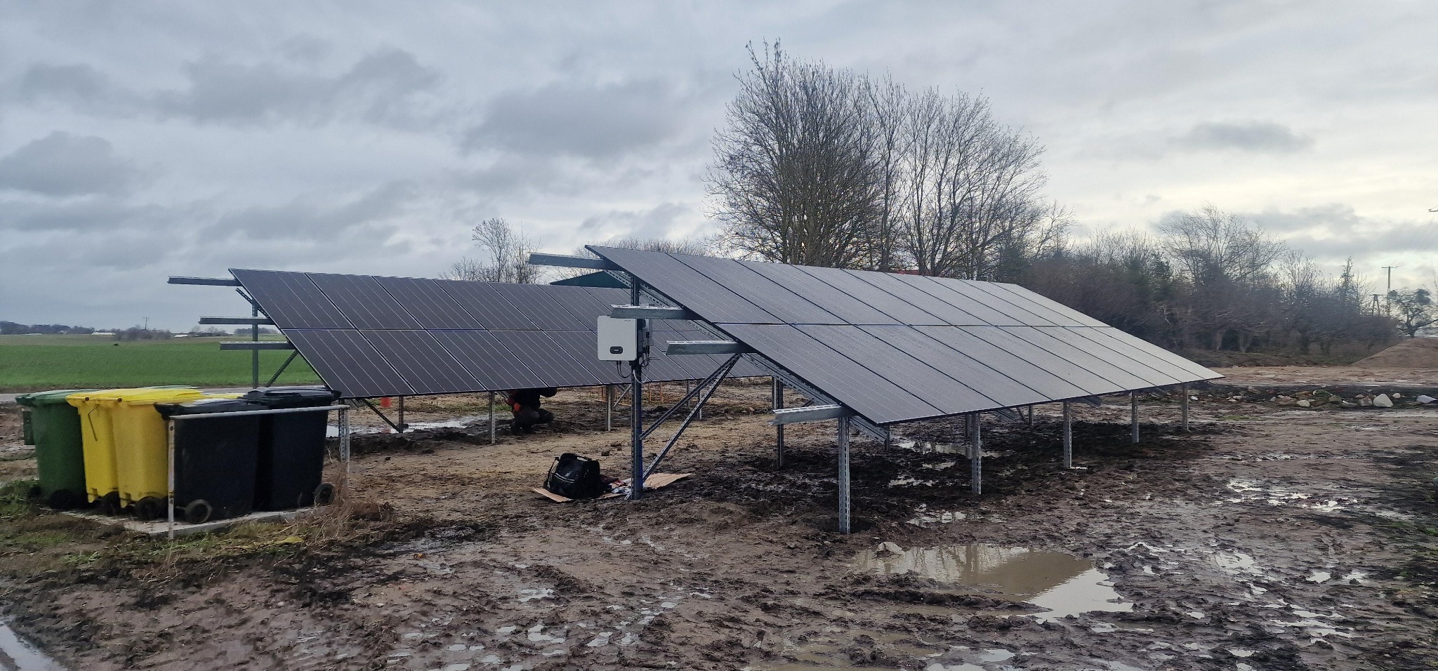 Montaż paneli fotowoltaicznych na metalowej konstrukcji w terenie. Widoczne panele, inwerter, narzędzia i błotniste podłoże. Pochmurne niebo w tle.