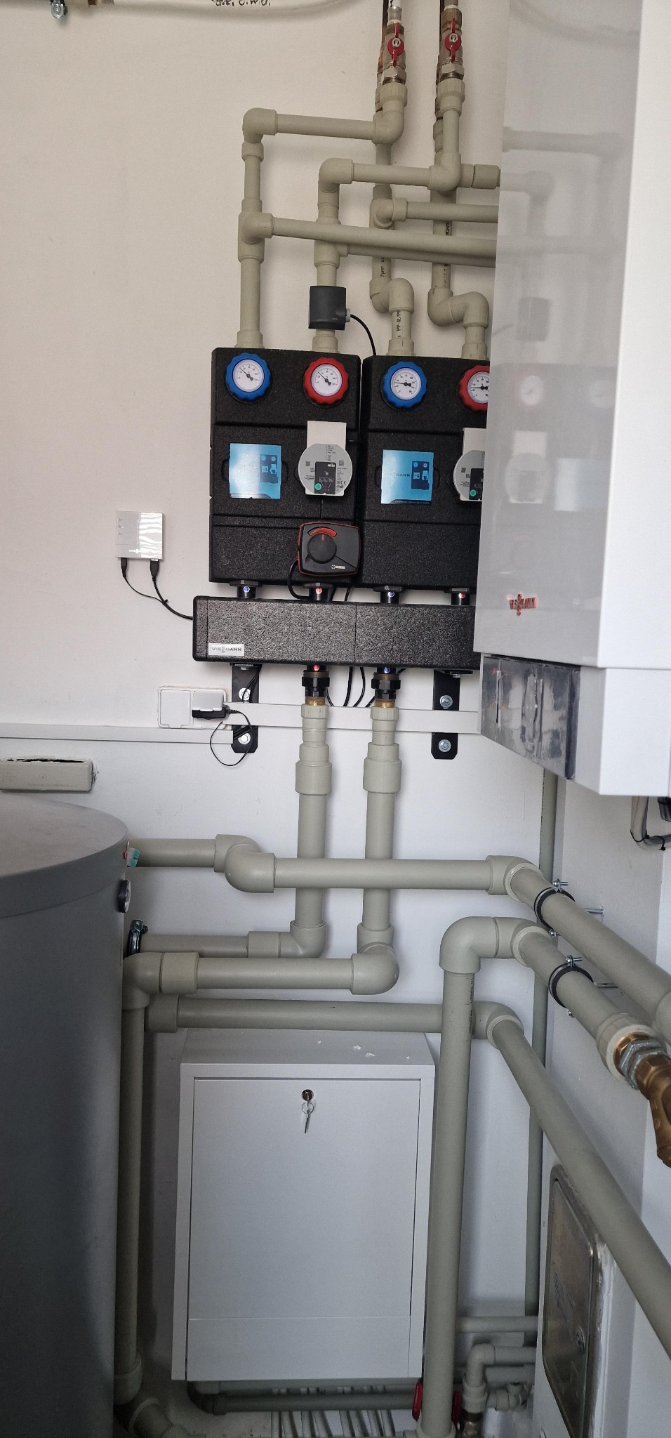 Węzeł cieplny z kotłem Viessmann, manometrami i licznymi rurami z tworzywa w jasnym pomieszczeniu technicznym. Widoczny zbiornik i szafka z kluczem.
