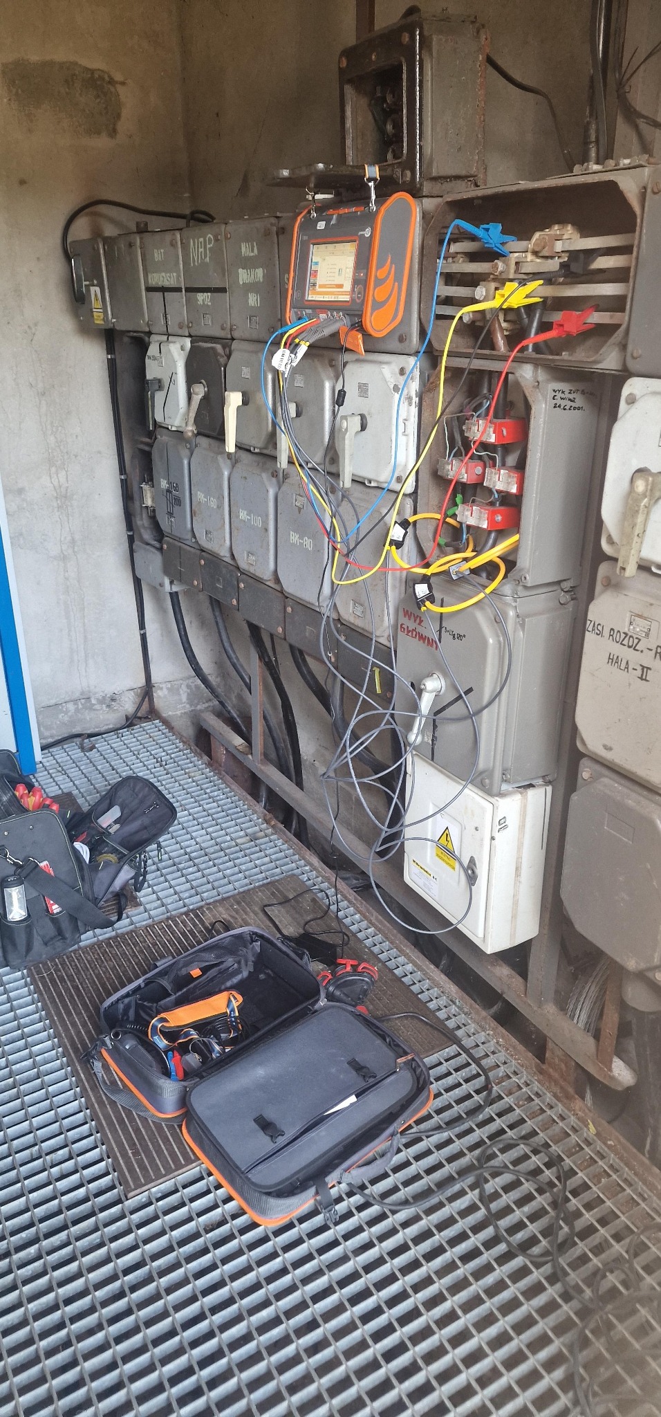 Szara skrzynka elektryczna z podłączonym miernikiem i przewodami. Otwarta torba narzędziowa na metalowej kratownicy. Pomiary elektryczne w hali przemysłowej.