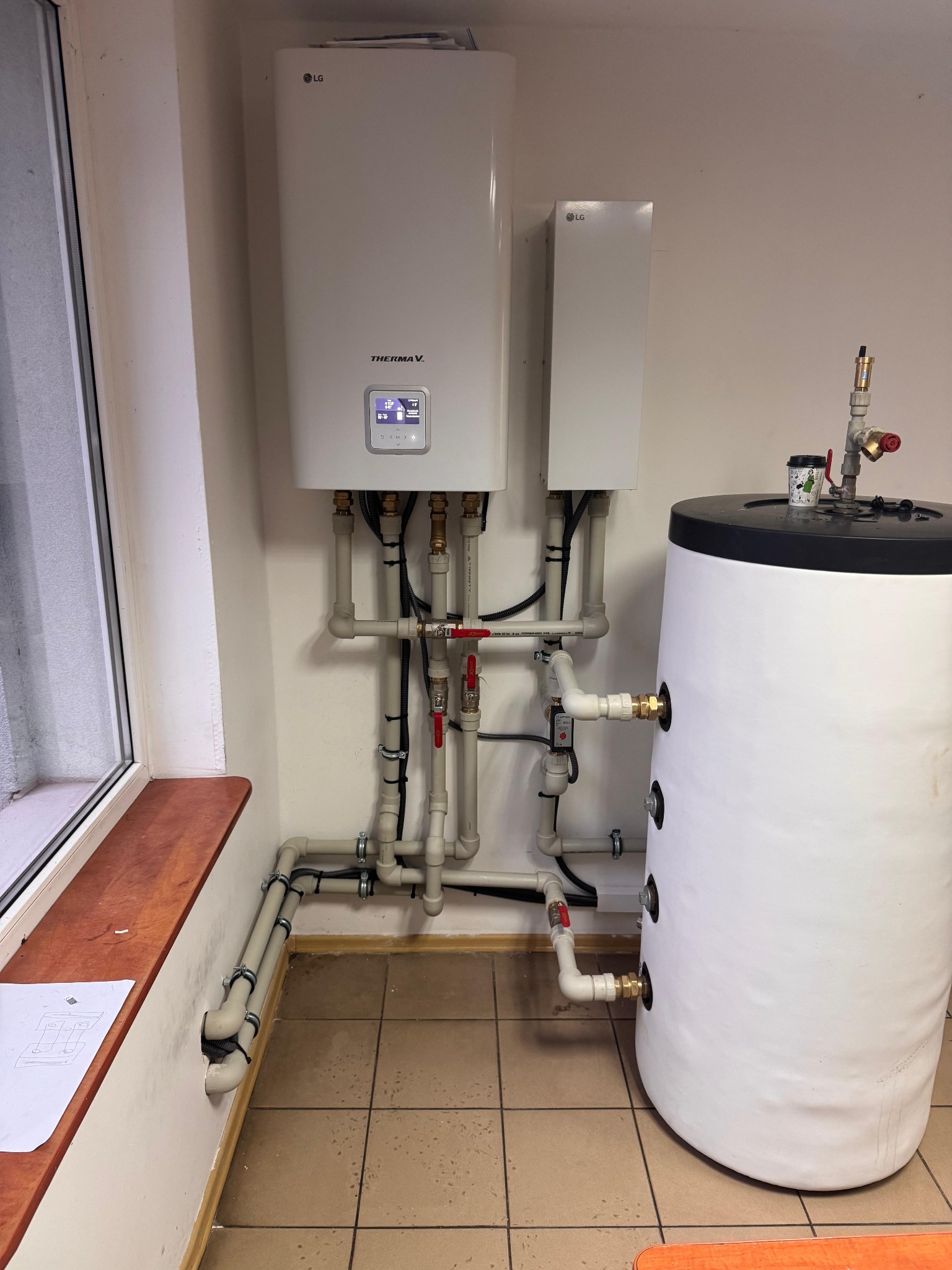 Biała pompa ciepła LG Therma V z zbiornikiem wody, połączona rurami z zaworami, zamontowana na ścianie w pomieszczeniu z oknem i brązowymi płytkami podłogowymi.