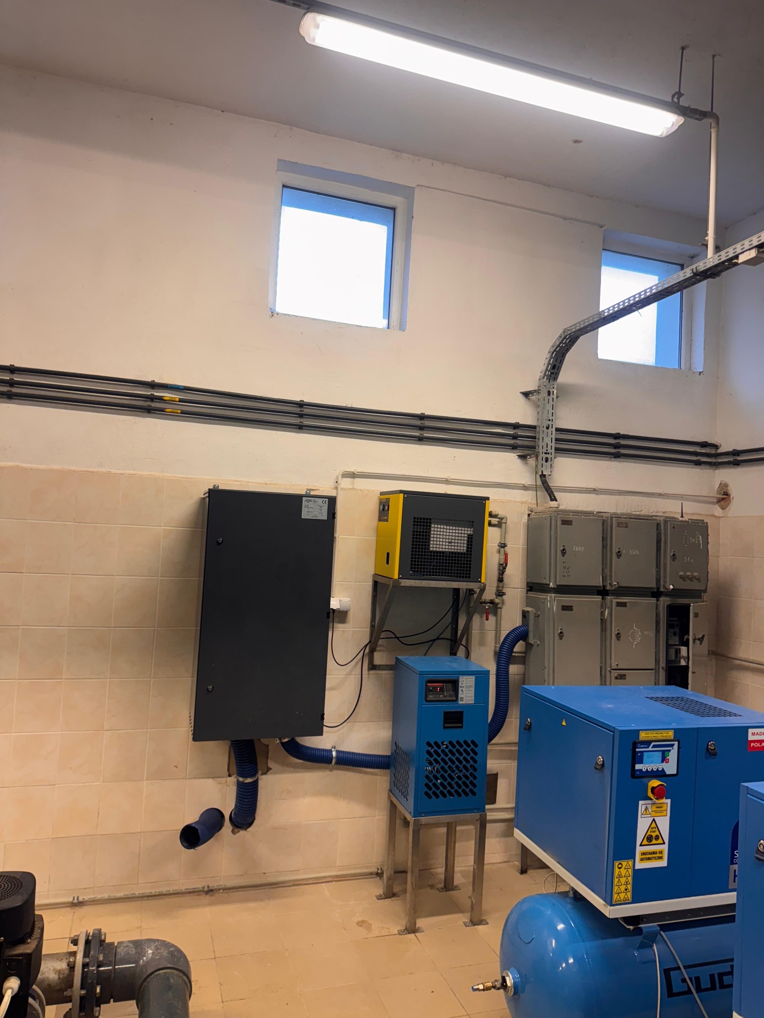 Kompresorownia: niebieskie i żółte kompresory, czarna szafa sterownicza na kafelkowanej ścianie. Widoczne rury i kable. Oświetlenie jarzeniowe.