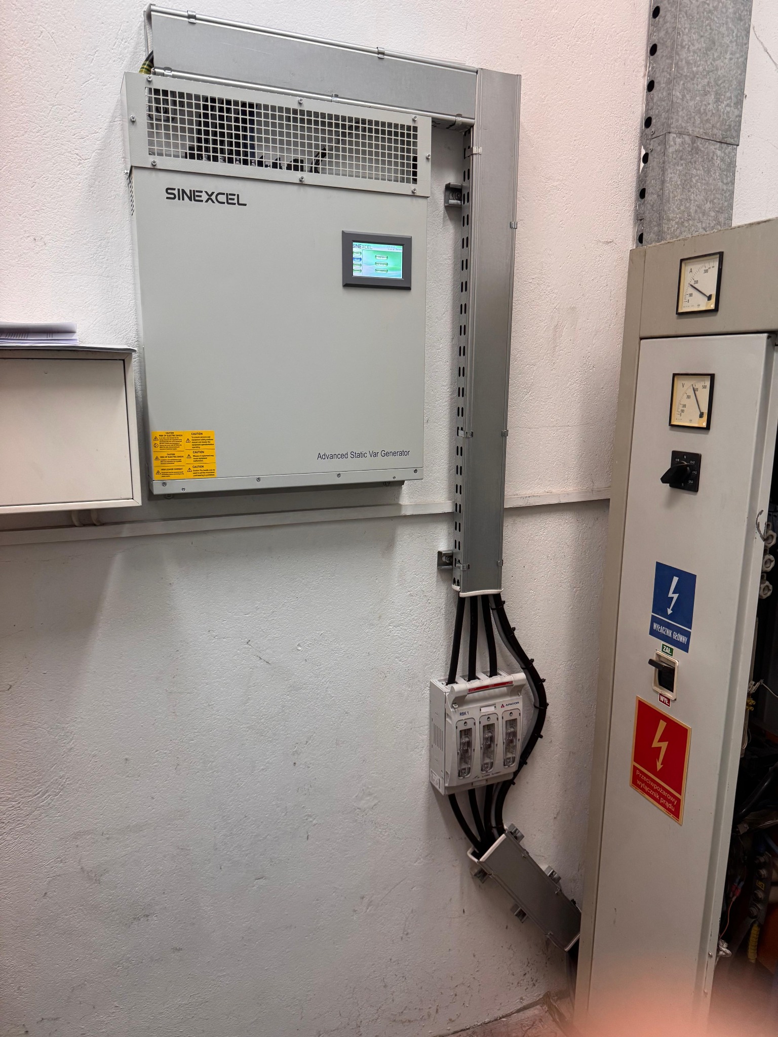 Szara szafa elektryczna Sinexcel z wyświetlaczem i okablowaniem na ścianie. Widoczne zabezpieczenia i oznaczenia ostrzegawcze. Ujęcie z dołu, oświetlenie naturalne.