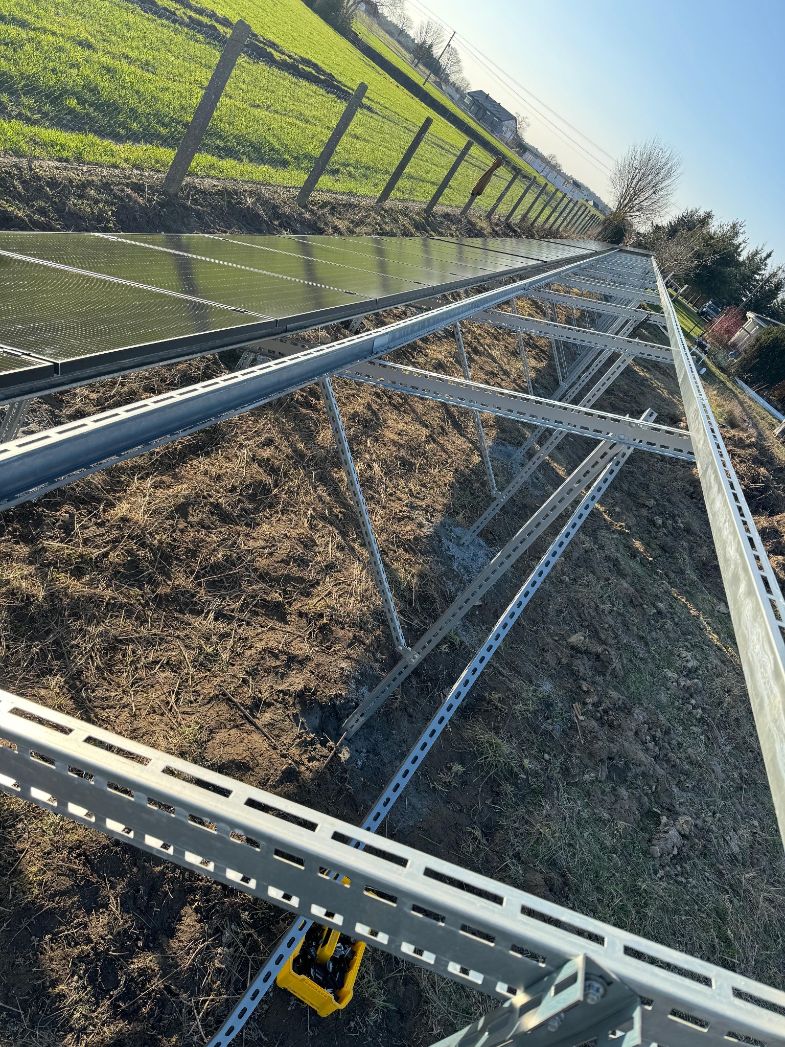 Montaż konstrukcji wsporczej pod panele fotowoltaiczne na gruncie. Widoczne metalowe profile i elementy mocujące. W tle zielone pole i ogrodzenie.