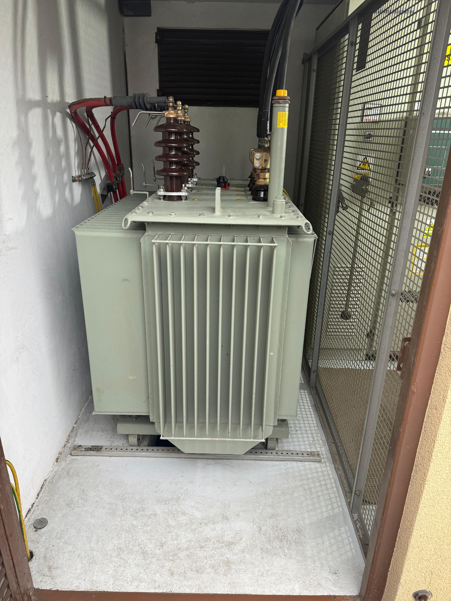 Transformator w obudowie z siatki, widoczne kable i izolatory. Element infrastruktury elektrycznej w pomieszczeniu technicznym.